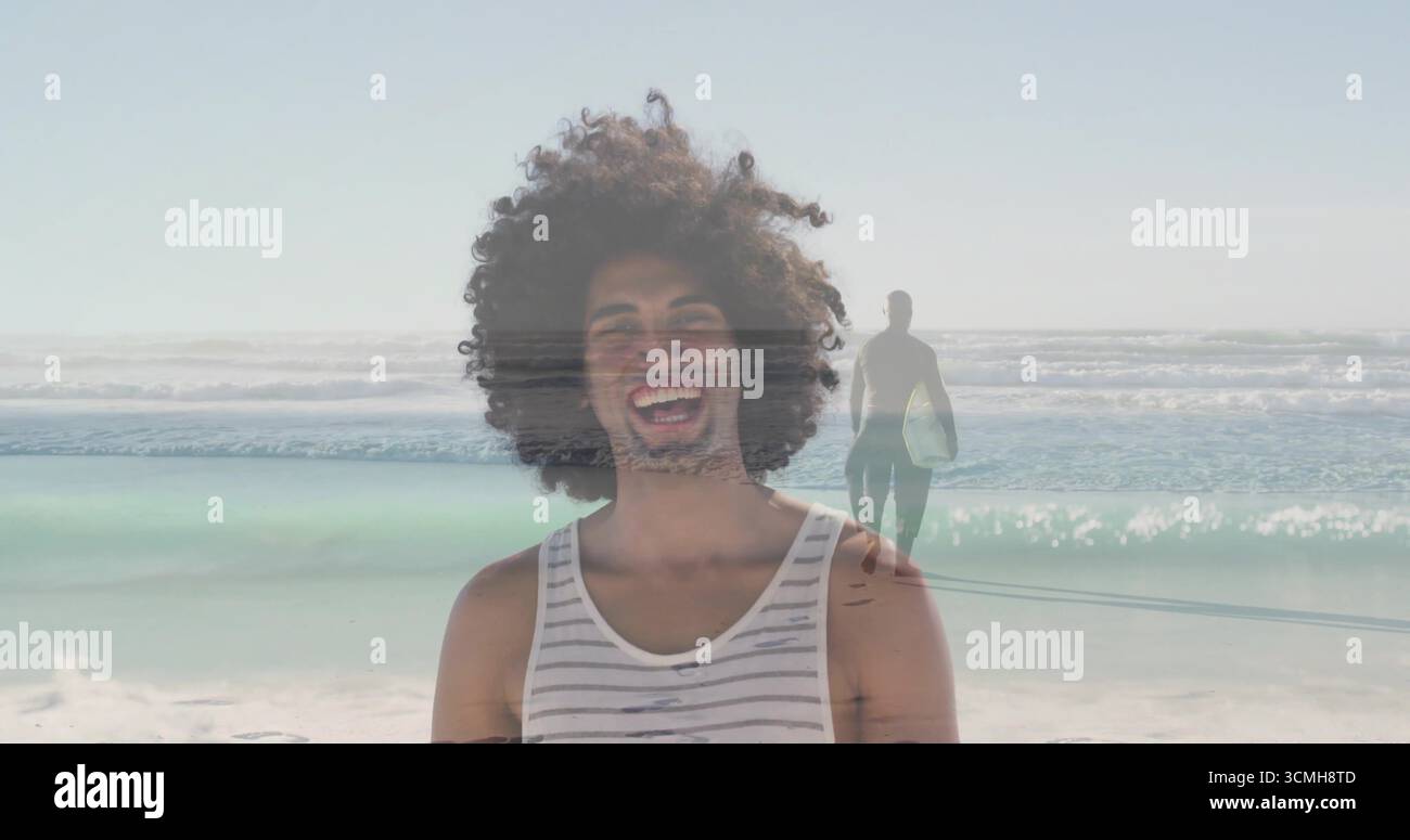Uomo sorridente che indossa una canotta a righe sulla spiaggia, con un secondo uomo che cammina nel surf che tiene il longboard Foto Stock