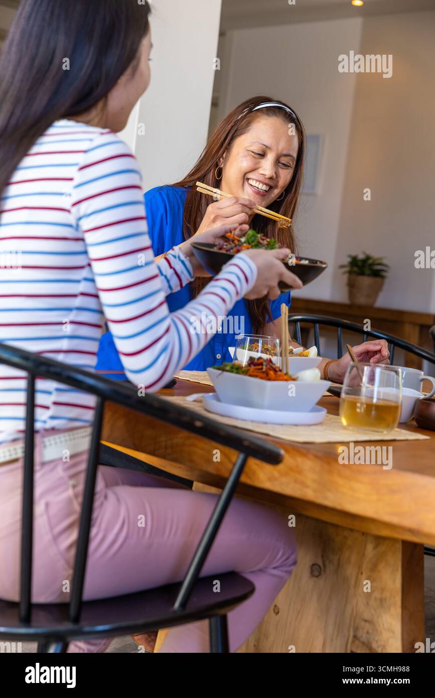 Madre e figlia asiatiche condividono l'insalata in ciotole nere usando bacchette al tavolo della sala da pranzo Foto Stock