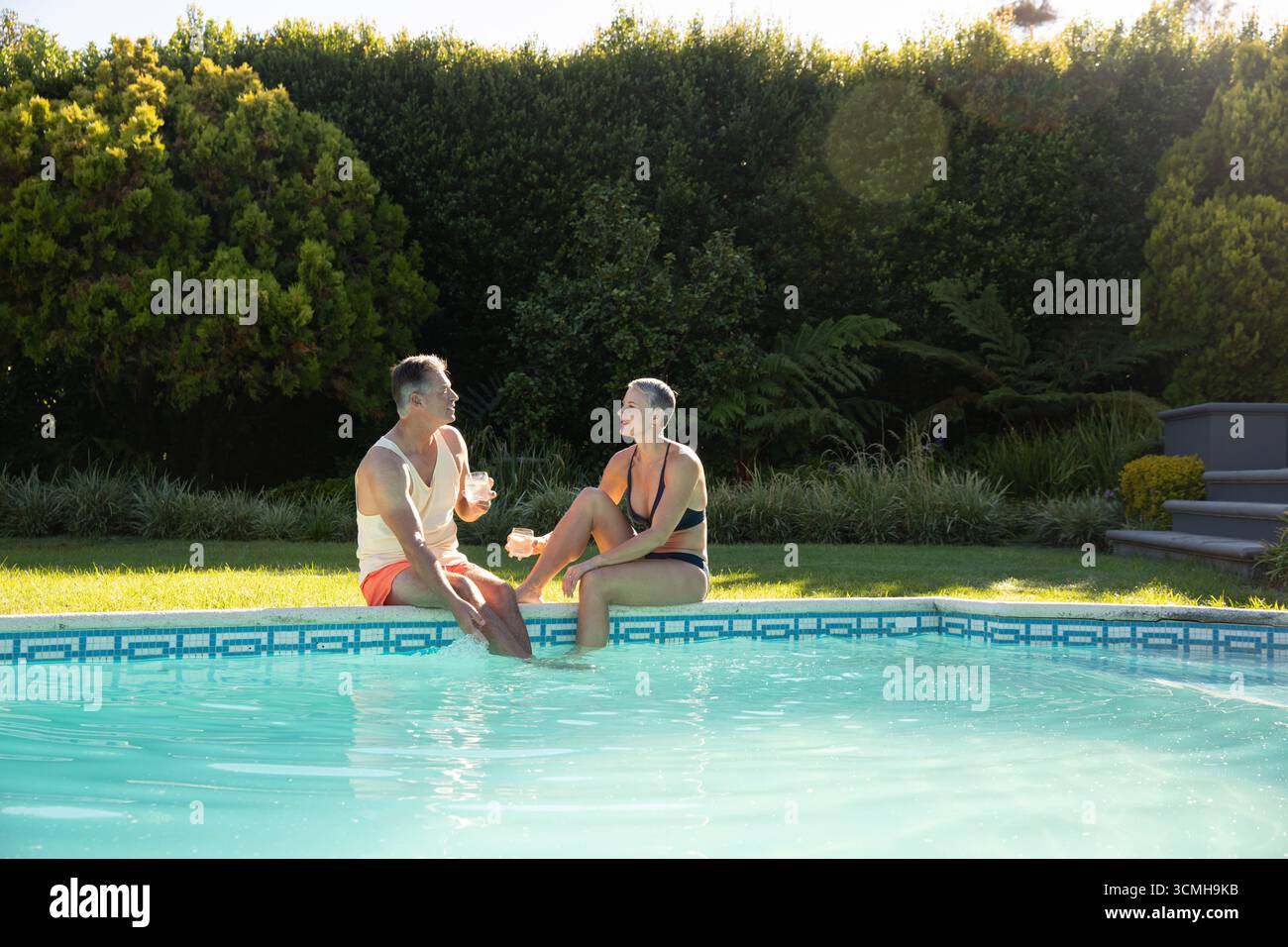 La coppia si siede a bordo piscina nel cortile sul retro, immergendo i piedi in acqua piastrellata mentre tiene le tazze Foto Stock