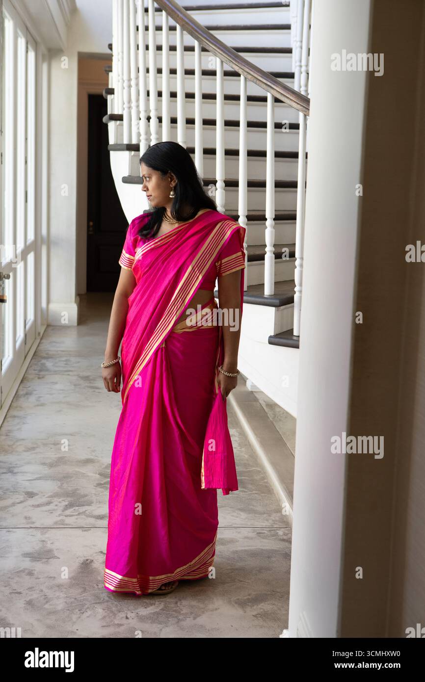 Donna indiana in piedi nel foyer, indossa vivaci sari rosa e gioielli d'oro accanto alle alte finestre Foto Stock