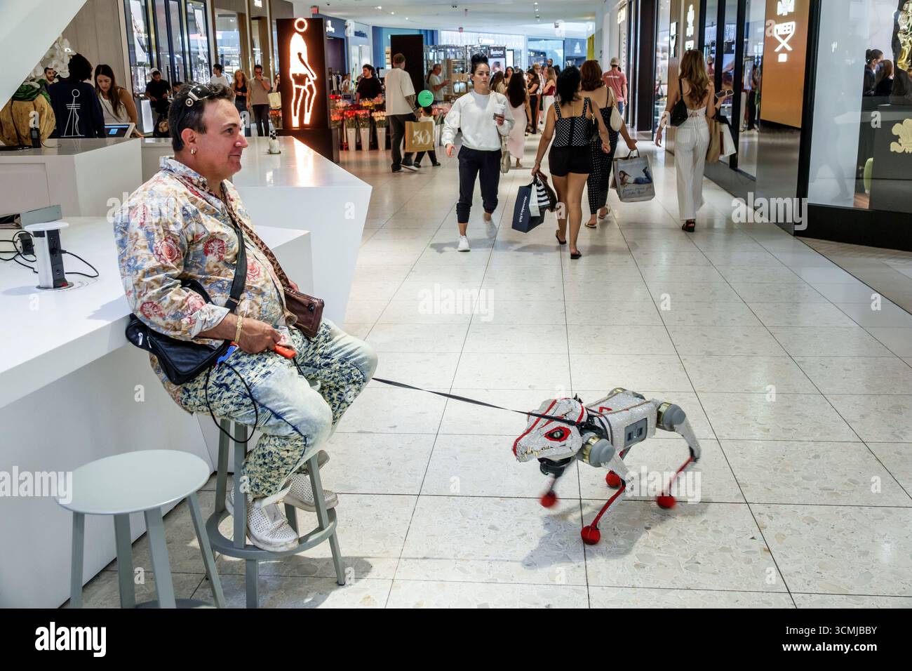 Centro commerciale Aventura Miami Florida, negozi interni, uomo seduto su uno sgabello con guinzaglio, cane robotico animatronico, intrattenimento tecnologico Foto Stock