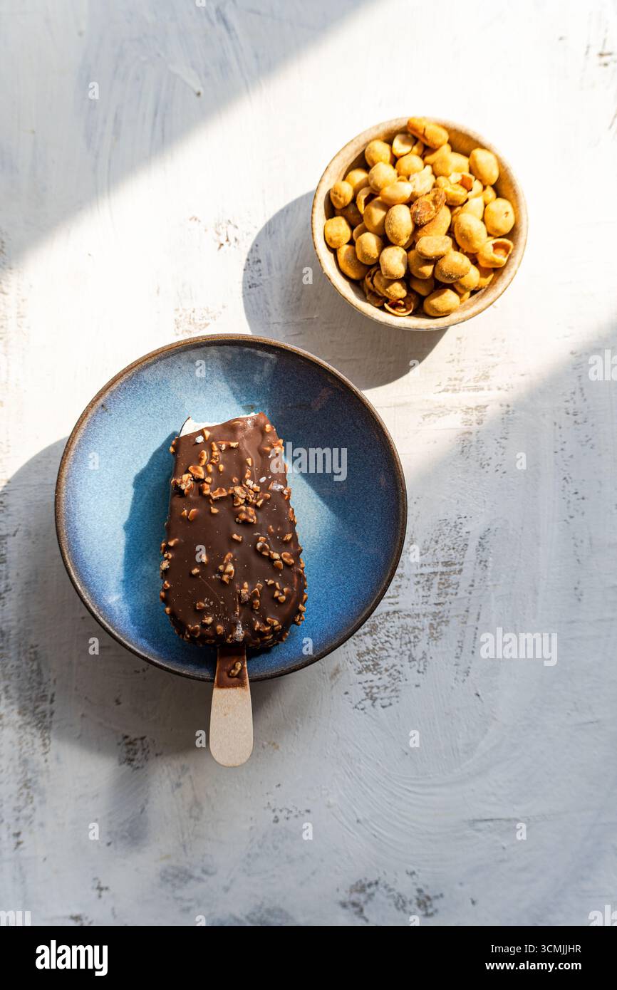 Vista dall'alto di un gelato ricoperto di cioccolato e noci e una ciotola di noci salate su un tavolo alla luce del sole Foto Stock