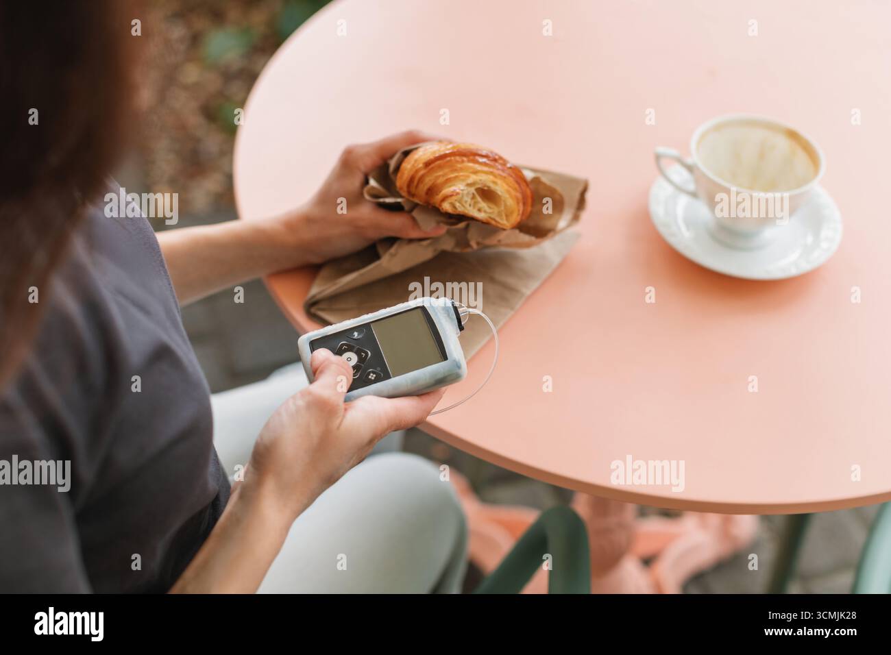 Primo piano di una giovane donna con diabete di tipo 1 seduta a un tavolino con un croissant e un caffè con una pompa per insulina Foto Stock
