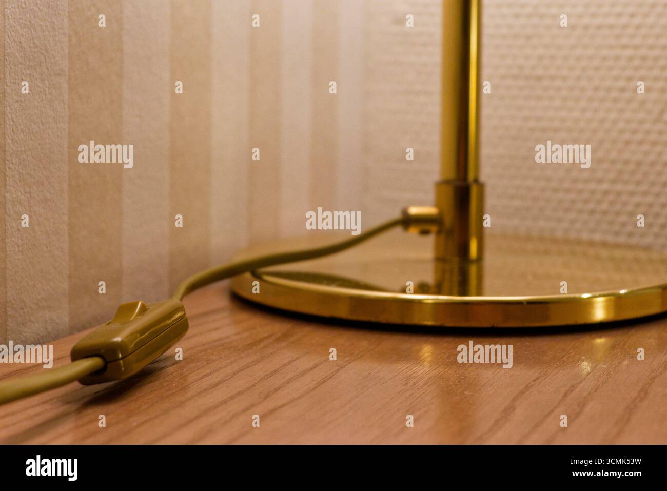 Una lampada da scrivania dorata poggia su un tavolo di legno accanto alla carta da parati a righe beige. Il cavo è dotato di un interruttore in linea e il paralume caldo diffonde una luce soffusa Foto Stock