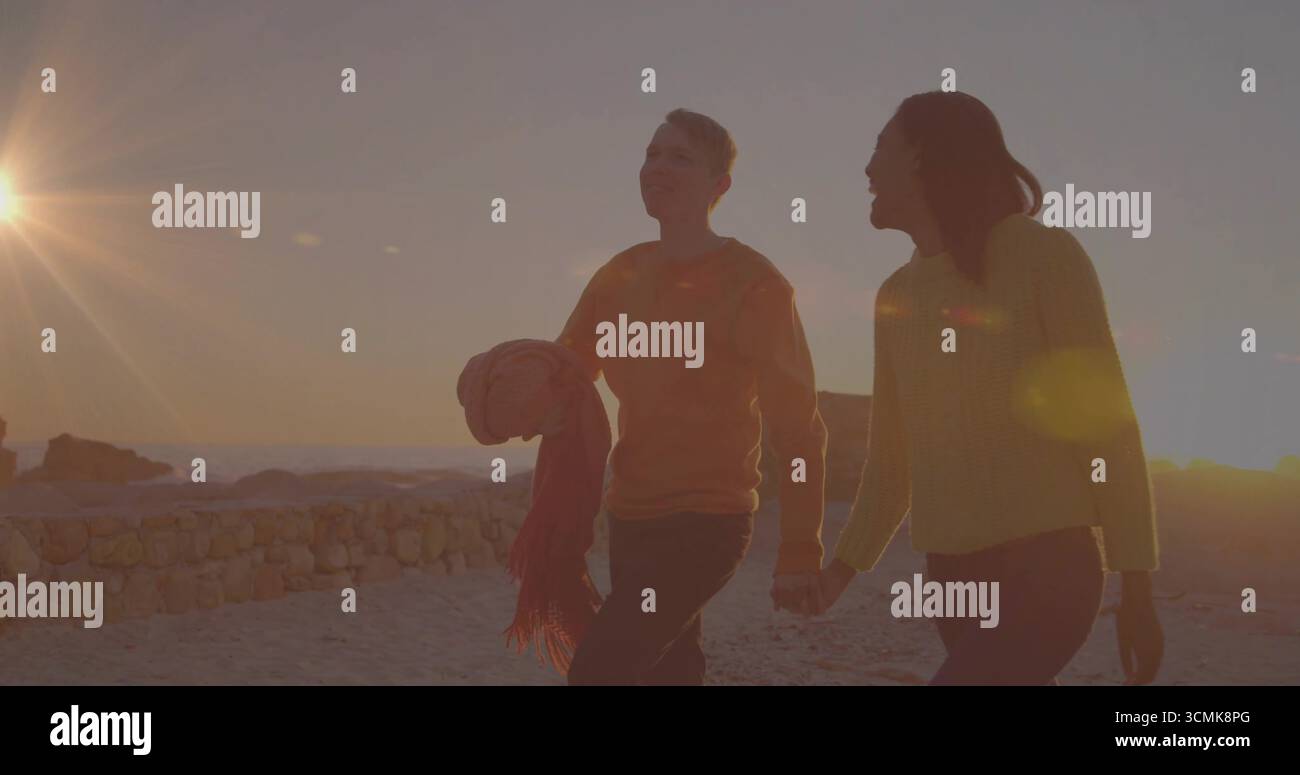 Coppia che cammina per mano lungo la spiaggia sabbiosa al tramonto, con coperta arrotolata e sciarpa con frange Foto Stock
