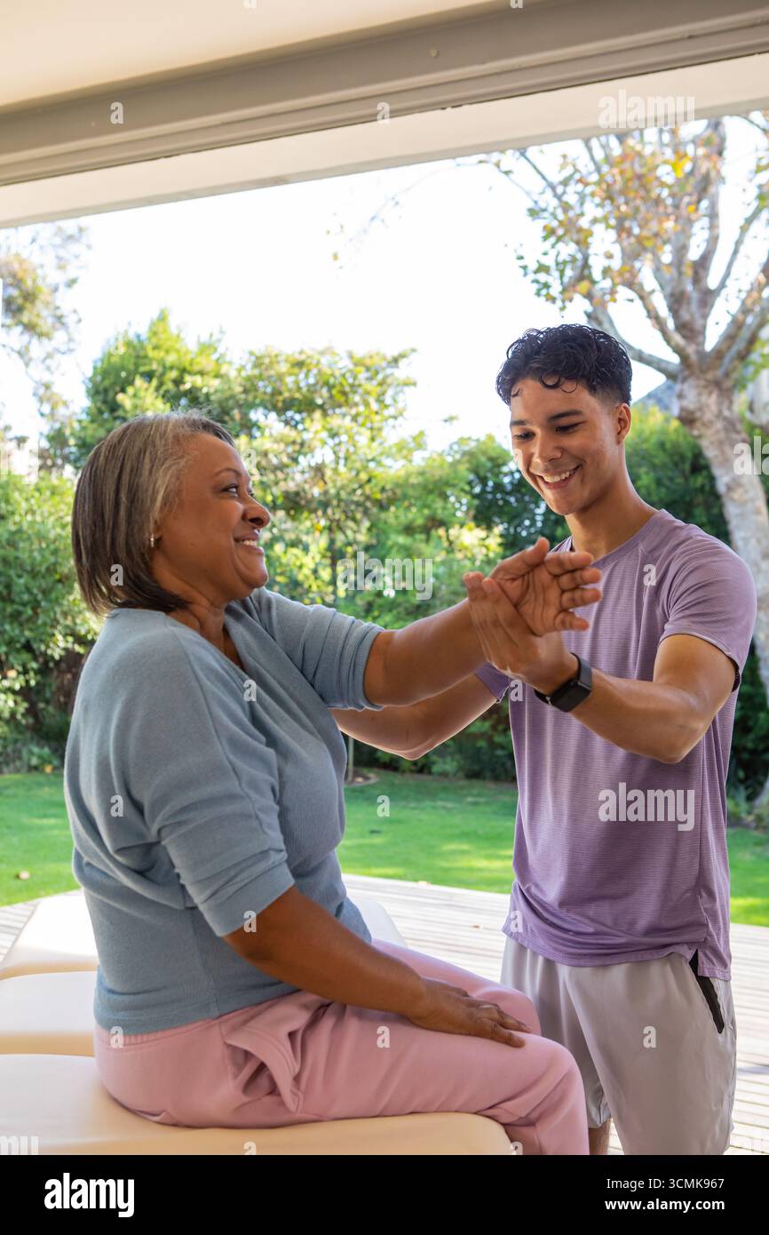 Terapeuta ispanica che guida una donna afroamericana anziana in braccio si allunga sul patio nel tavolo terapeutico Foto Stock
