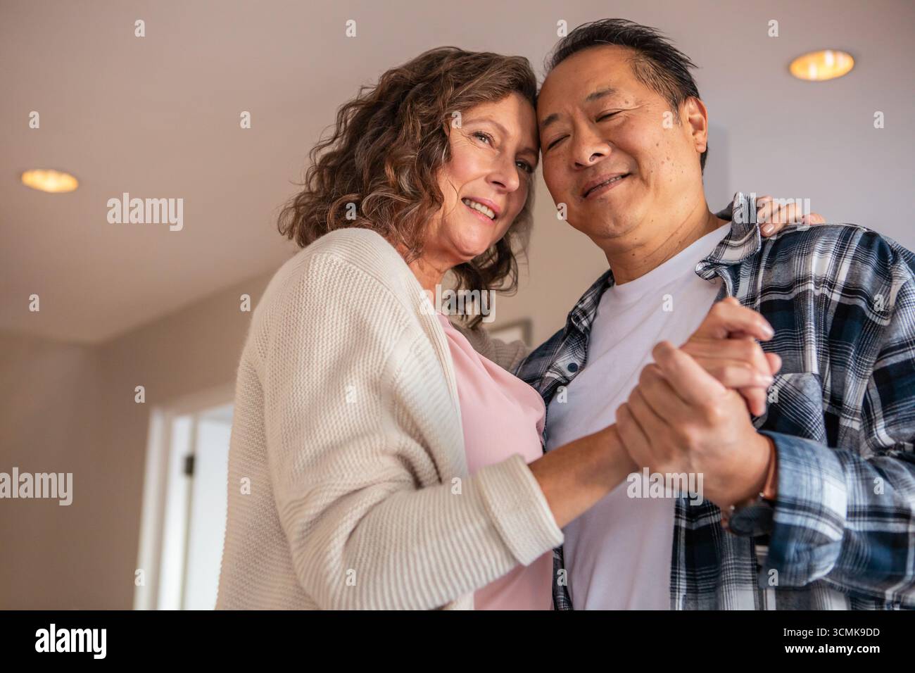 Coppia senior che balla nel soggiorno da pareti beige sotto le calde luci del soffitto che si tengono per mano Foto Stock