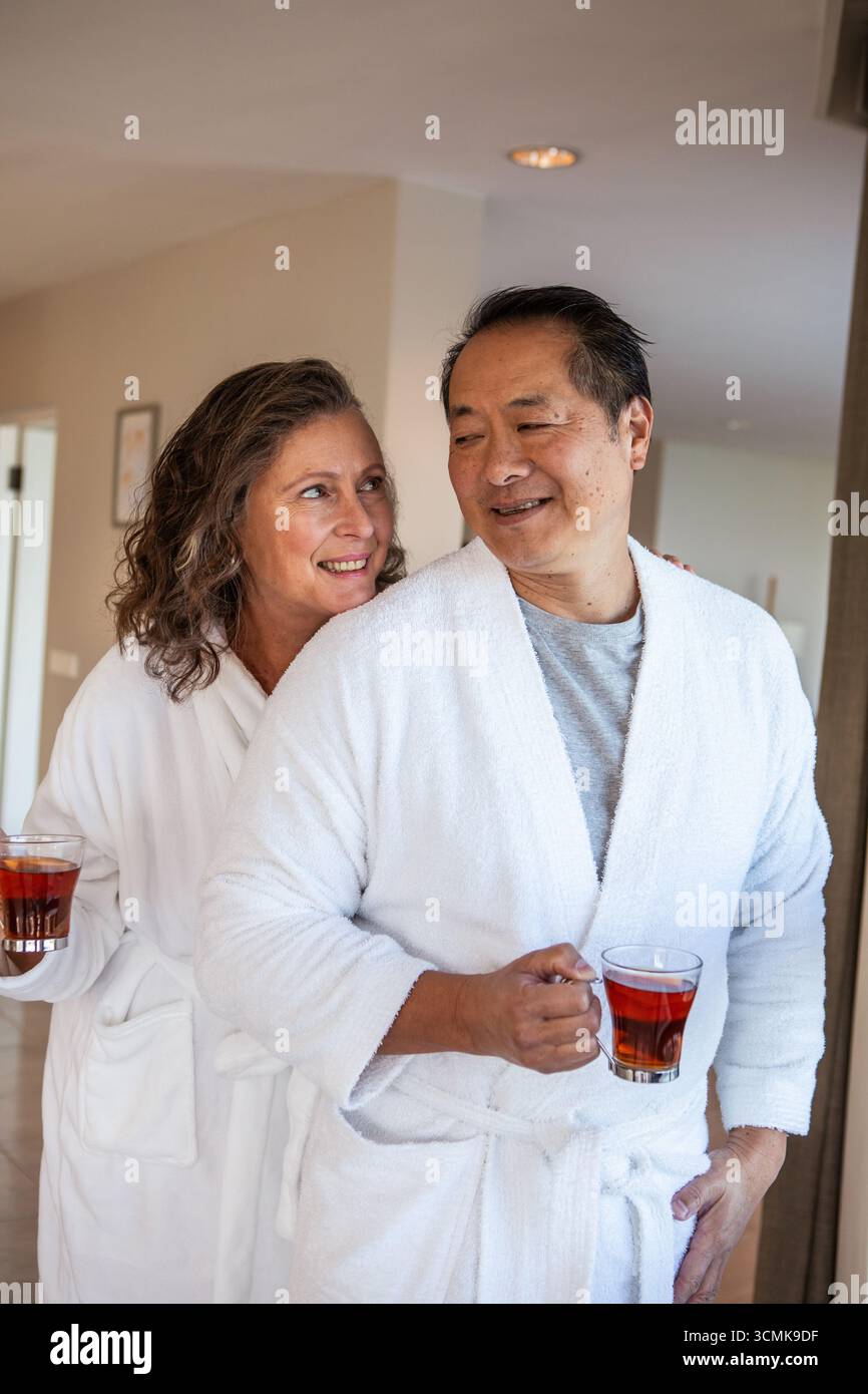 Diverse coppie in piedi in un corridoio morbido e casalingo con accappatoi bianchi che tengono tazze da tè rosse Foto Stock