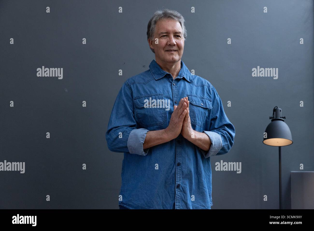 Uomo anziano in piedi in studio che indossa camicia in denim e pantaloni scuri con piantana Foto Stock