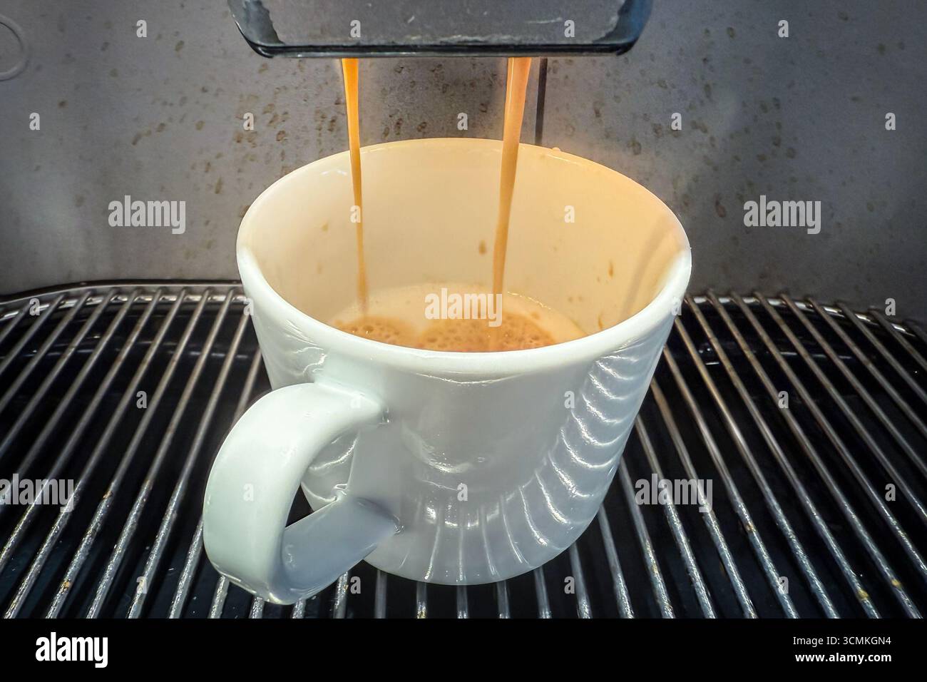 Vista ravvicinata di una tazza bianca riempita di caffè espresso da una macchina da chicco a una tazza, due flussi versano sulla crema cremosa sopra un vassoio antigoccia di metallo, un au Foto Stock