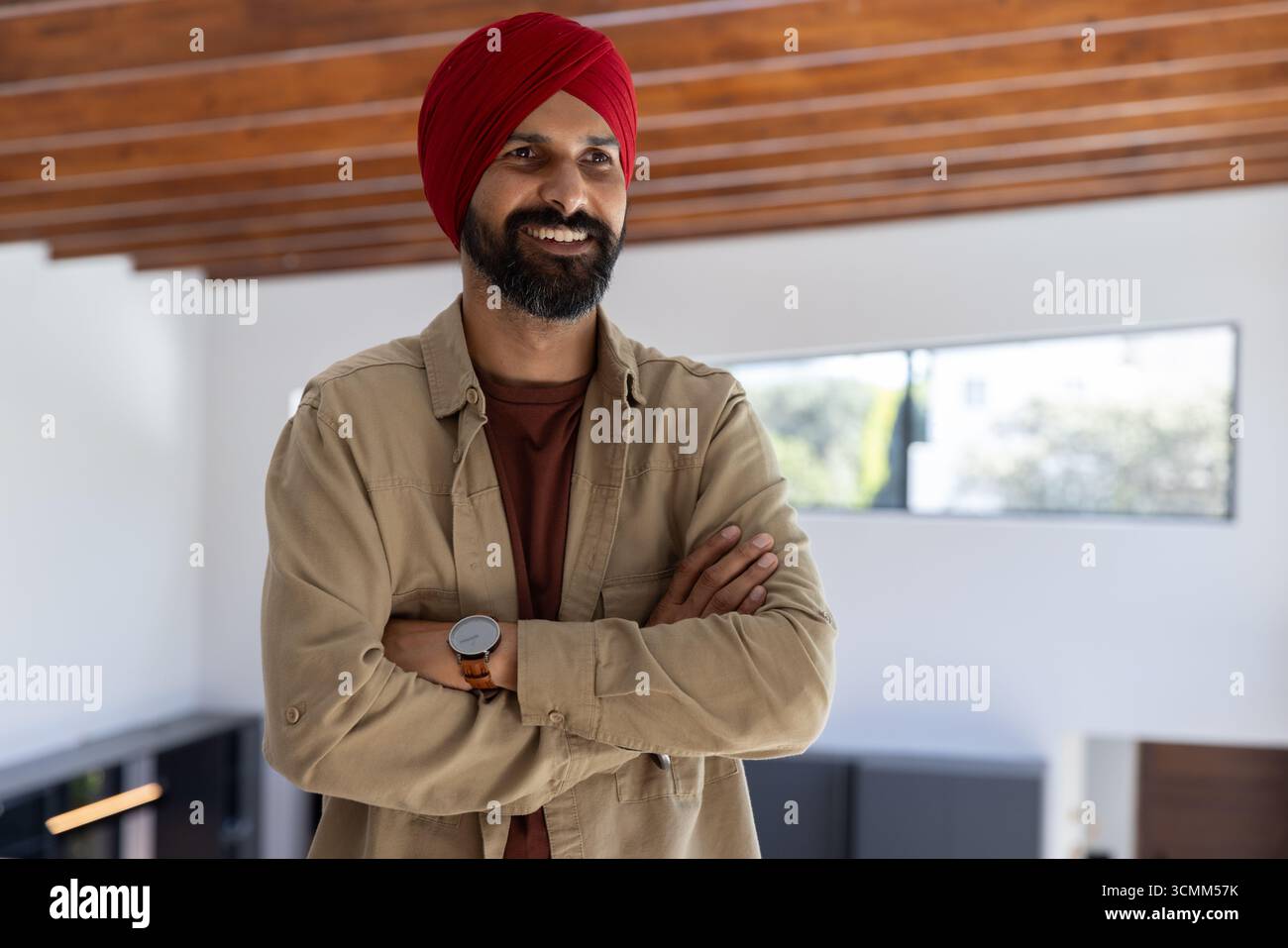 Uomo indiano medio adulto che incrocia le braccia mentre sorride a casa con orologio da polso e travi in legno Foto Stock