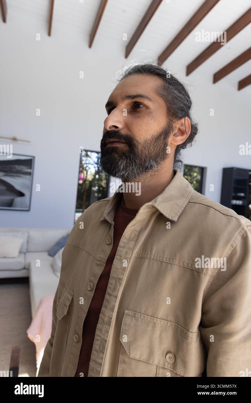 Uomo indiano medio adulto che indossa una camicia beige e una t-shirt ruggine in piedi nel soggiorno accanto al divano Foto Stock