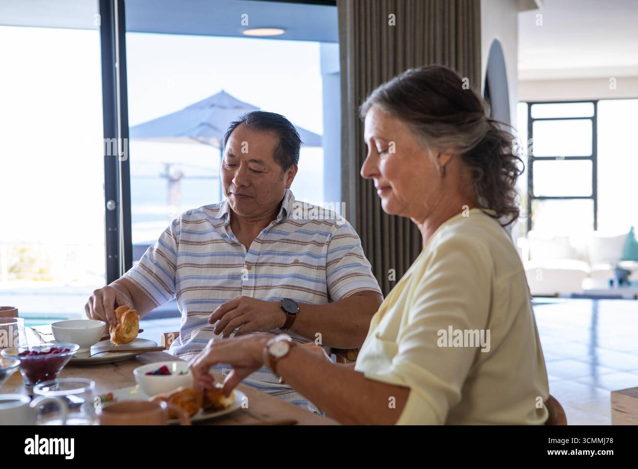 La coppia senior si siede accanto a un tavolo da pranzo in legno a casa, assaggiando croissant e vasetti di marmellata Foto Stock