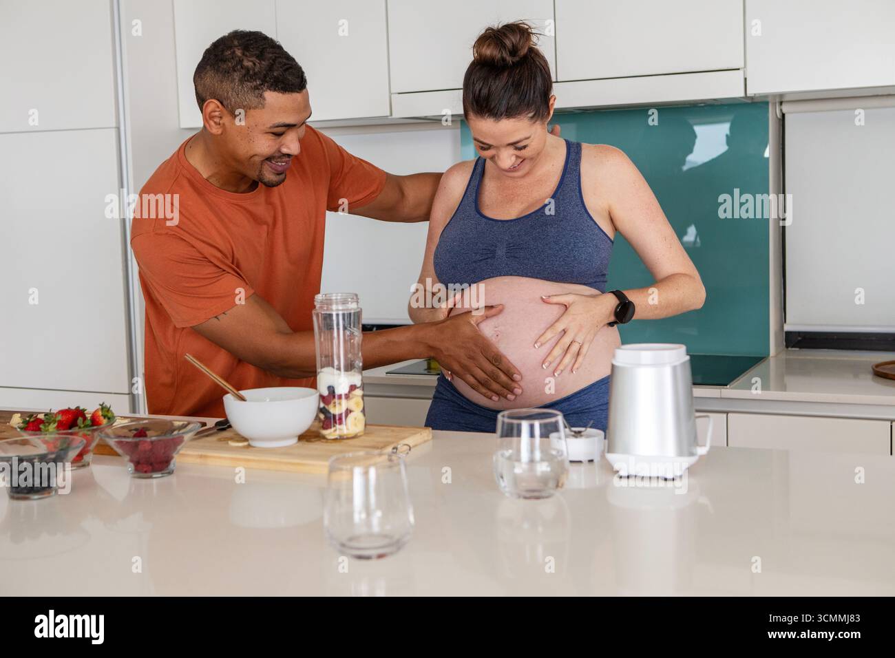 Diverse coppie che si aspettano di mettere le mani sulla pancia incinta nella cucina di casa con ciotole e frullatore Foto Stock