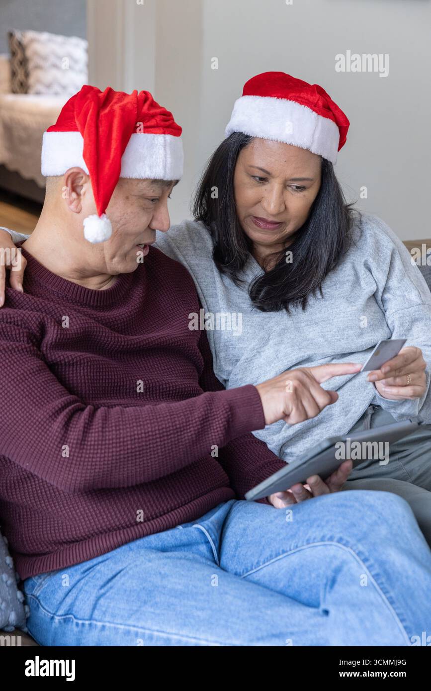 Coppia senior in cappelli di babbo natale che naviga online su tablet con carta di credito in salotto Foto Stock