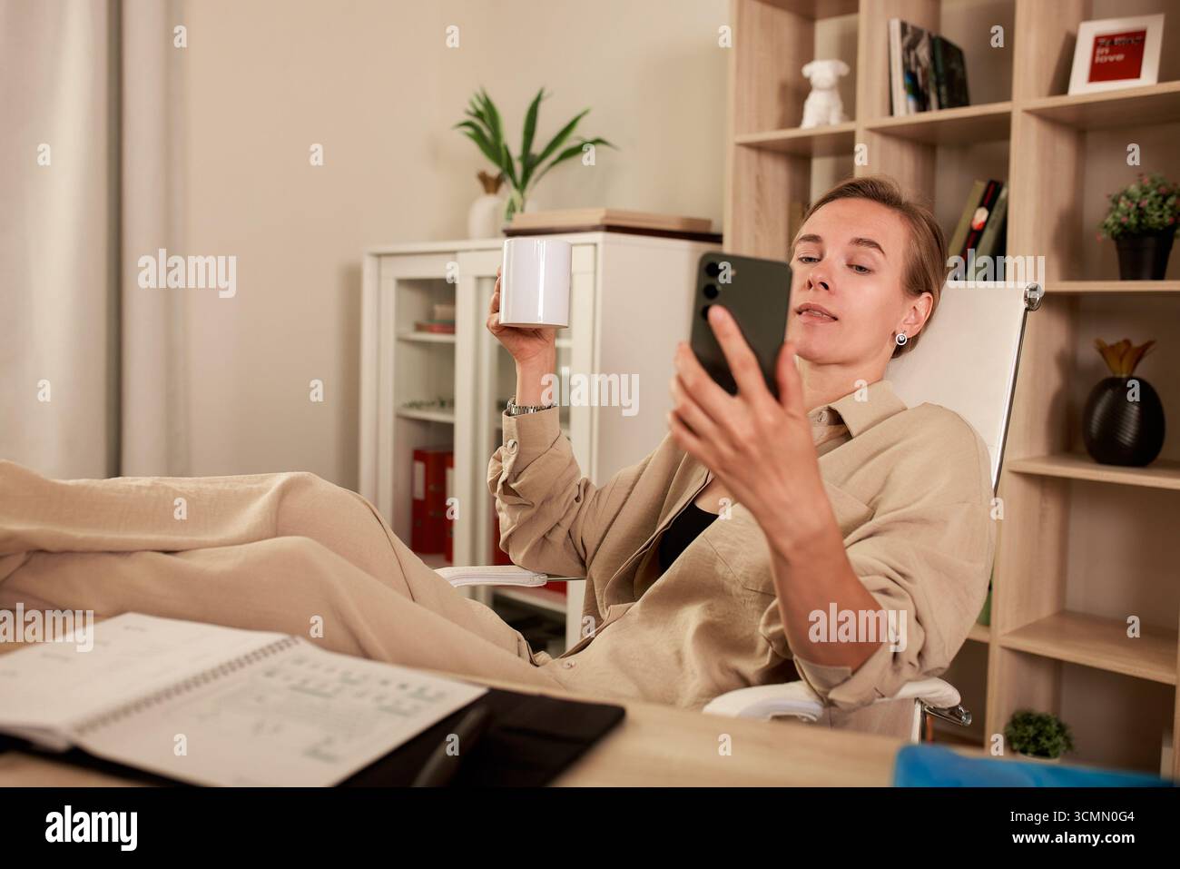 Un professionista è seduto alla propria scrivania in una postura rilassante, tenendo in mano una tazza di caffè. Guardano il loro smartphone in un accogliente ufficio domestico Foto Stock