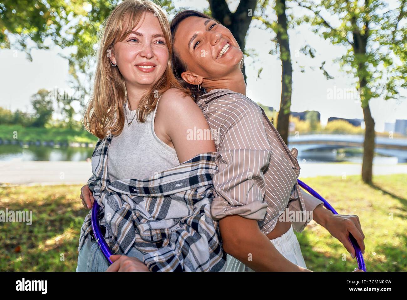 In una giornata di sole, due donne si divertono a fare attività all'aperto sorridendo e tenendo in mano i cerchi hula. Si trovano in un parco circondato da alberi e da un fiume sereno, radiati Foto Stock