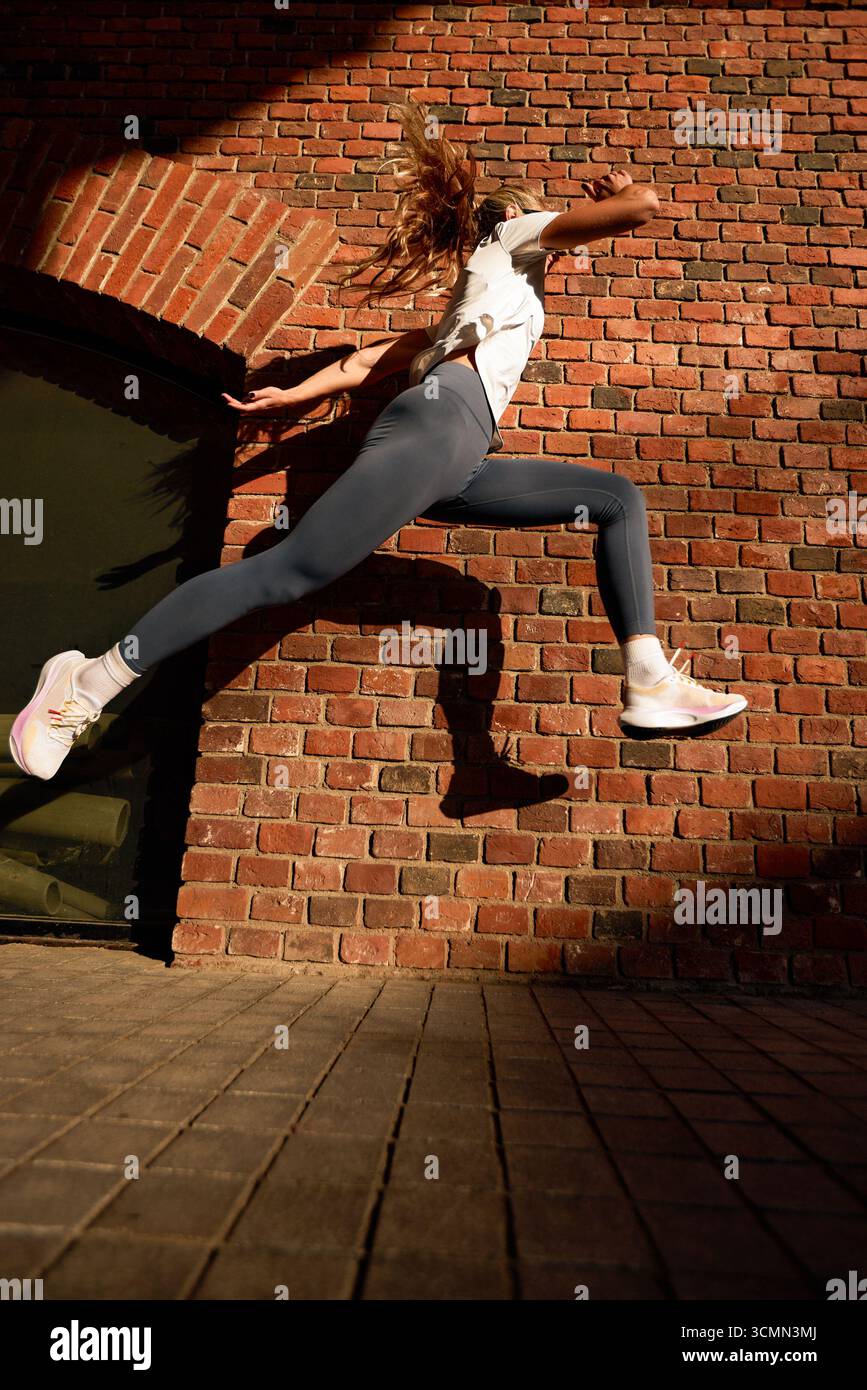 Una donna atletica salta dinamicamente con energia sullo sfondo di una parete di mattoni testurizzati, con la luce del sole che proietta ombre definite. L'immagine CA Foto Stock