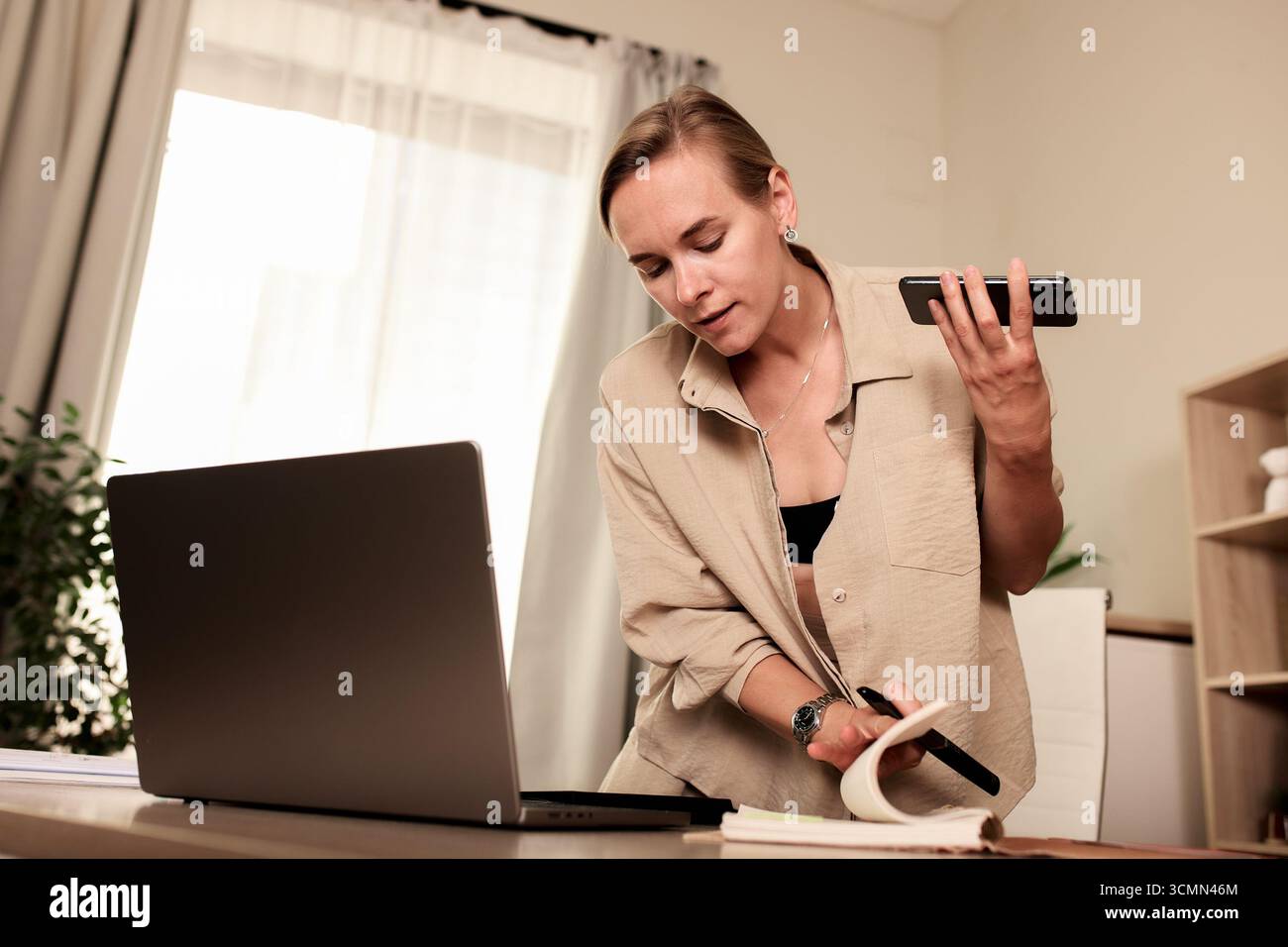 Una donna professionista che lavora alla sua scrivania, multitasking utilizzando uno smartphone, un notebook e prendendo appunti. L'immagine ritrae uno spazio di lavoro moderno, Highl Foto Stock
