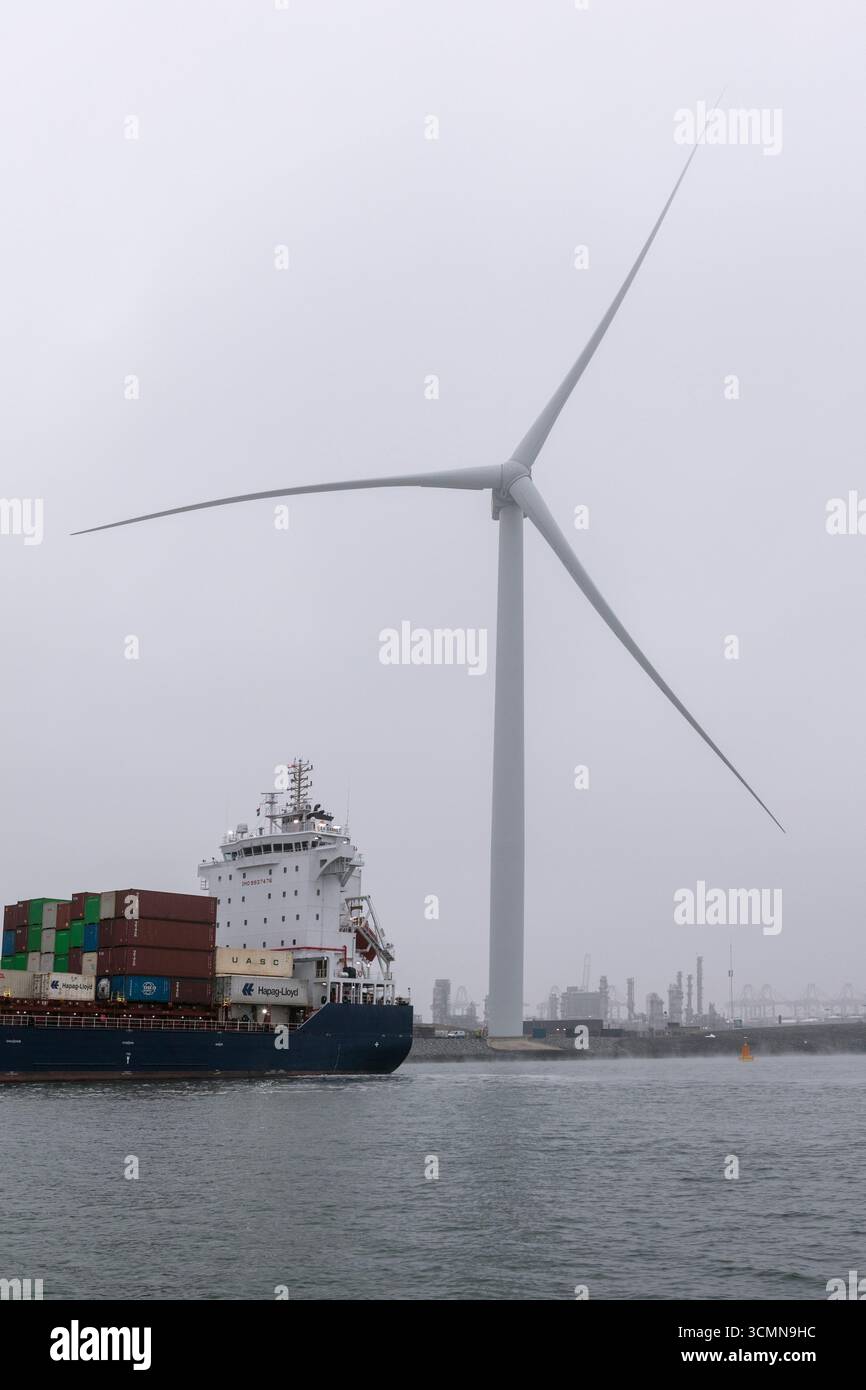 La nave portacontainer EA Gannet è ormeggiata in una giornata nebbiosa nel porto di Rotterdam, Paesi Bassi, con una turbina eolica che si innalza sullo sfondo sopra il lungomare industriale e acque calme. Foto Stock