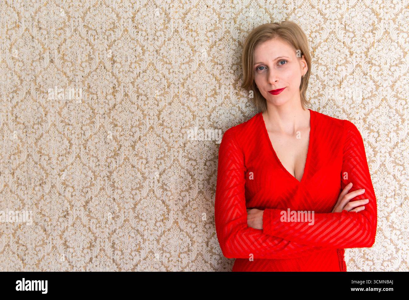 Tilburg, Nethetlands. La musica classica insegnante che indossa un abito rosso appoggiata contro una lettura della parete della camera. Foto Stock