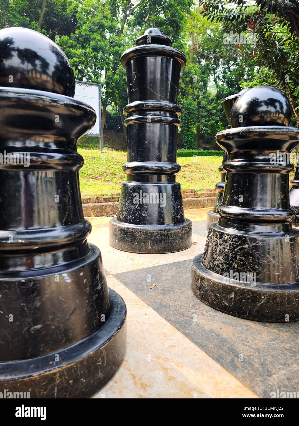 Un gigantesco pezzo da re nero è incorniciato da altri pezzi da scacchi all'aperto posizionati su una grande scacchiera con sfondo naturale. Foto Stock