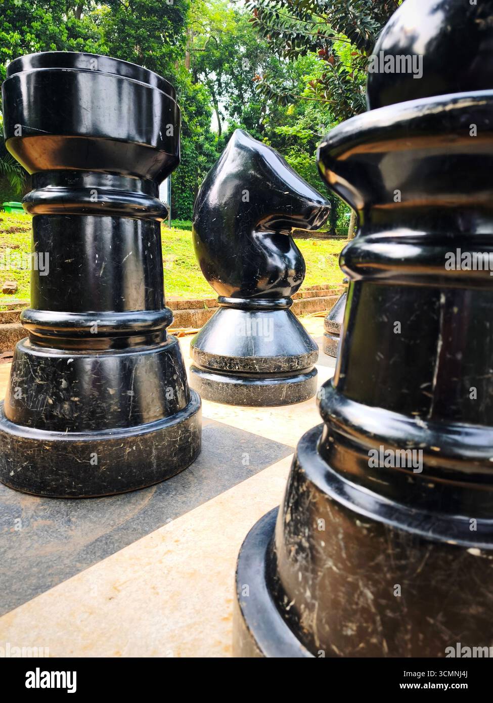 Un gigantesco pezzo di cavaliere nero si trova tra il pedone e i pezzi di scacchi disposti su una grande scacchiera all'aperto. Foto Stock