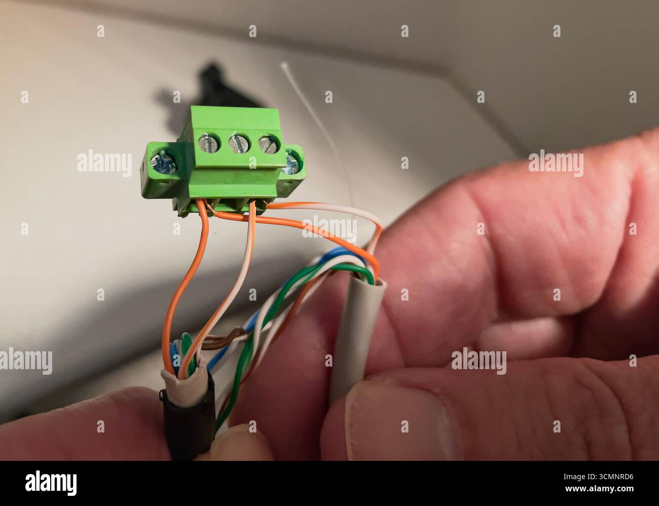 Primo piano di una mano che tiene una morsettiera verde con tre fili collegati (arancione, bianco e verde), che emergono da un cavo grigio, contro una spia B. Foto Stock