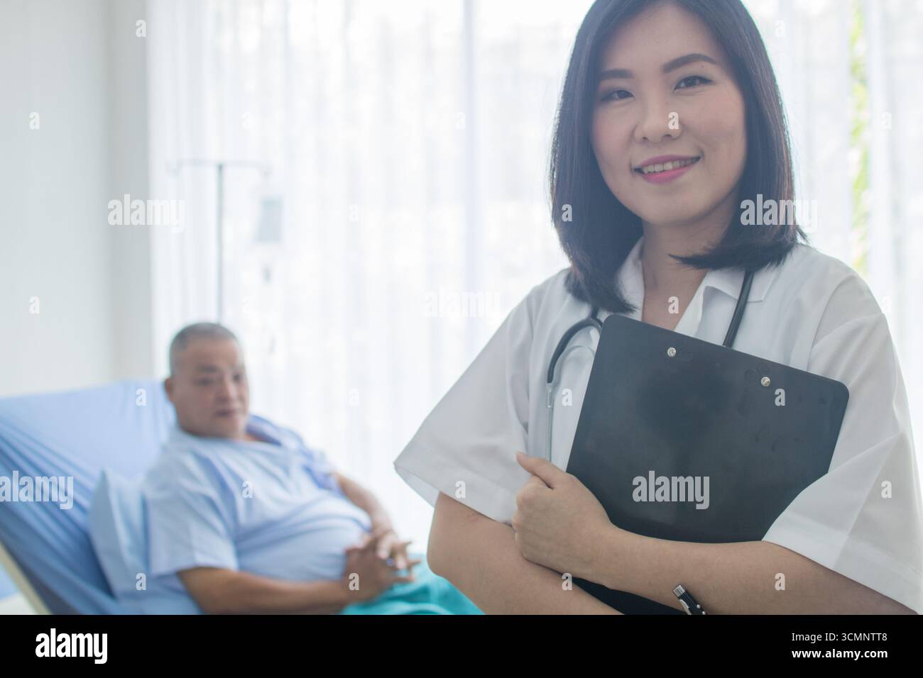 Una giovane dottoressa tiene il fascicolo medico di un paziente. C'è una paziente anziana sdraiata sul letto dietro di lei. Foto Stock
