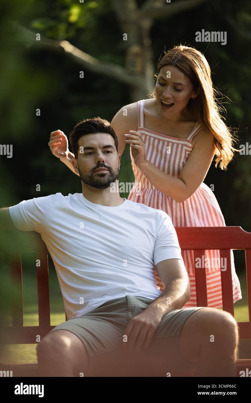 Donna gioiosa in piedi dietro la panchina il suo fidanzato indifeso è seduto, pronto a coprirsi gli occhi e a sorprenderlo Foto Stock