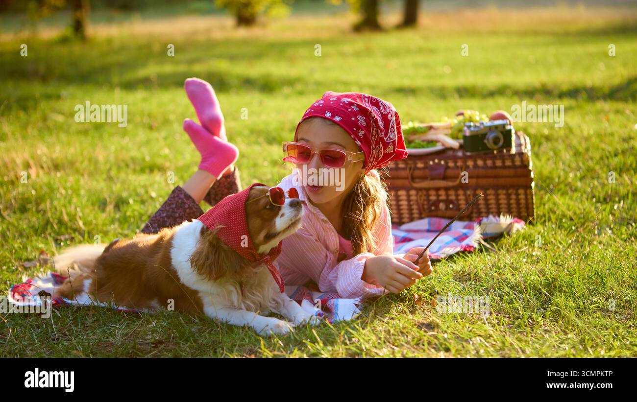 Ragazza in bandana rossa sdraiata su una coperta con il cane in occhiali da sole godendosi un picnic Foto Stock