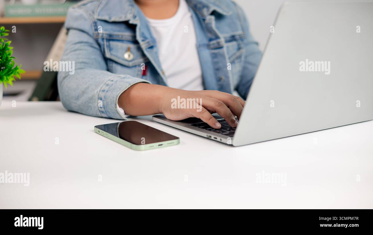 Primo piano di un uomo che utilizza un computer portatile con smartphone sulla scrivania, lavoro freelance, ufficio remoto, comunicazione digitale e moderno stile di vita aziendale per te online Foto Stock