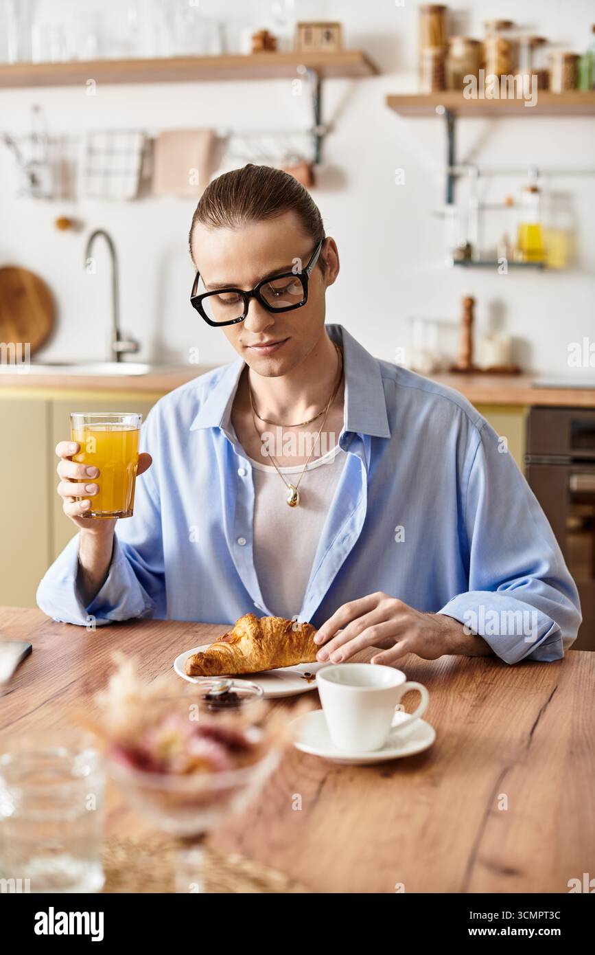 Un giovane e bello uomo dallo stile femminile si siede su un tavolo di legno, assaporando la colazione. Foto Stock