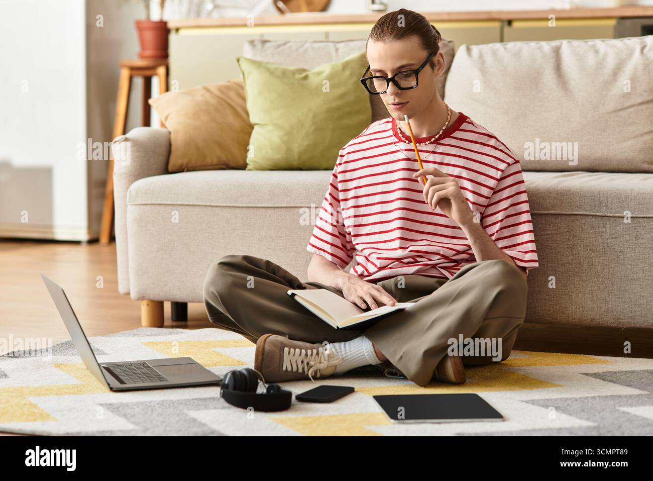 Un giovane dallo stile femminile si siede sul pavimento, pensando profondamente mentre scrive su un quaderno. Foto Stock