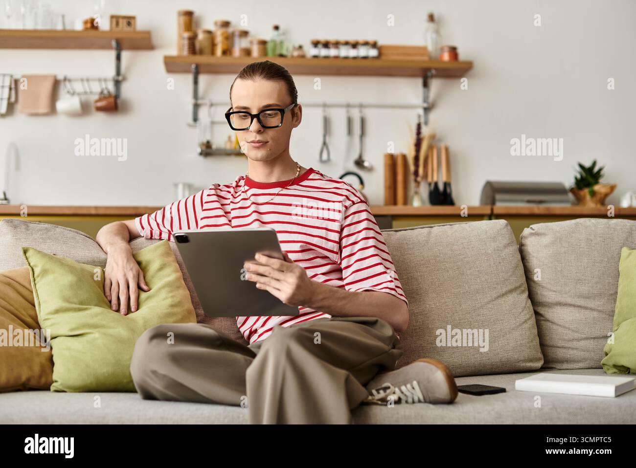 Un giovane uomo rilassato dallo stile femminile esplora i contenuti sul suo tablet in un ambiente accogliente. Foto Stock