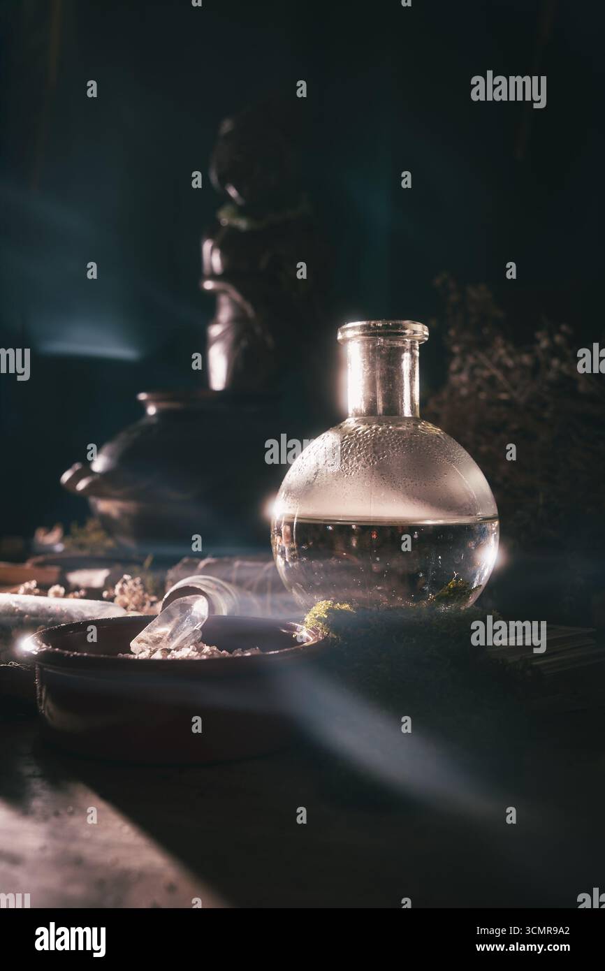 Misteriosa bottiglia di pozione in vetro su un altare Wiccan poco illuminato con erbe, muschio, cristalli ed elementi rituali che brillano in una luce mistica soffusa. Foto Stock