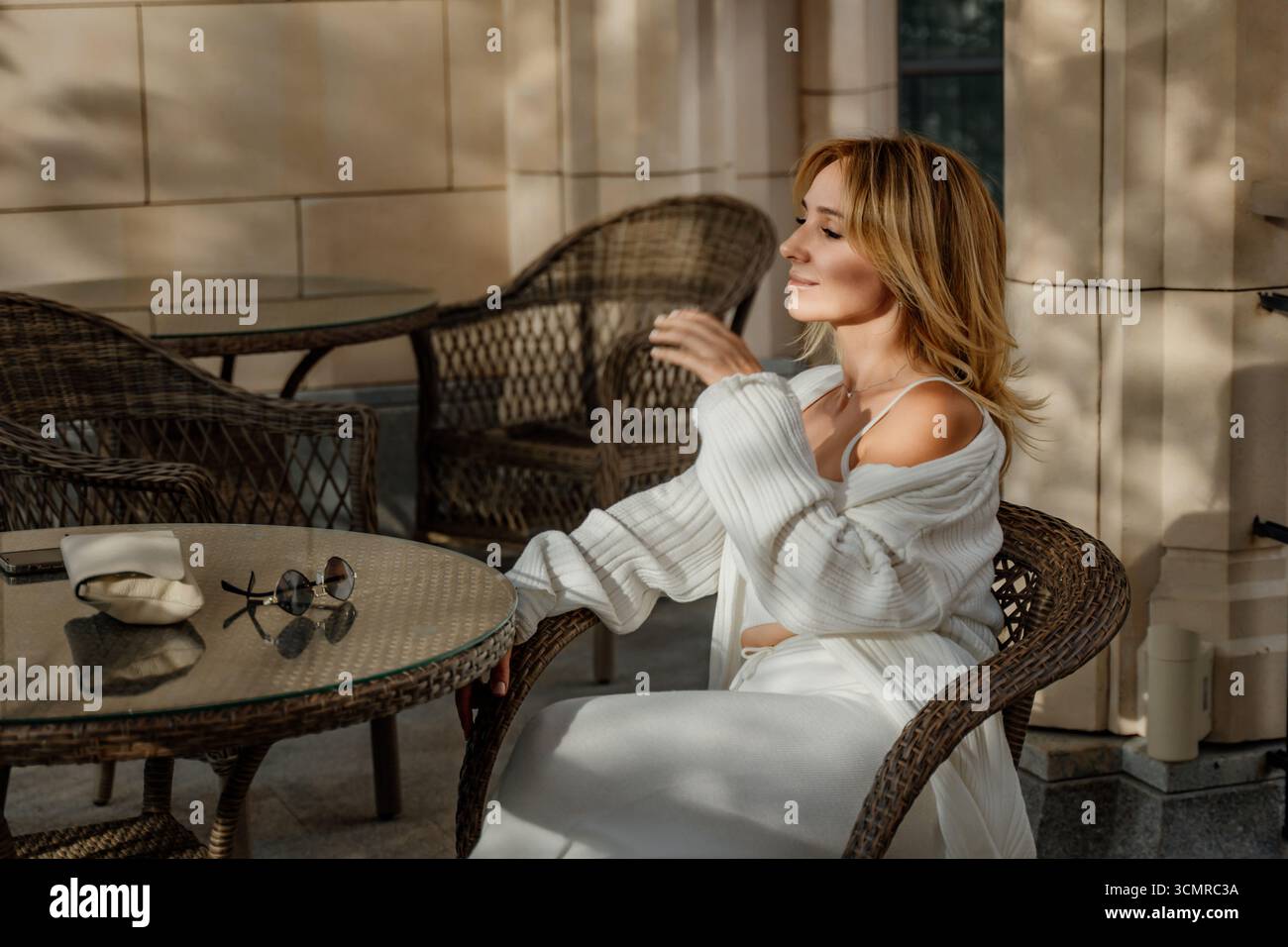 Woman Robe Patio - donna con accappatoio bianco seduta su una sedia da patio con un bicchiere d'acqua e occhiali da sole. Foto Stock