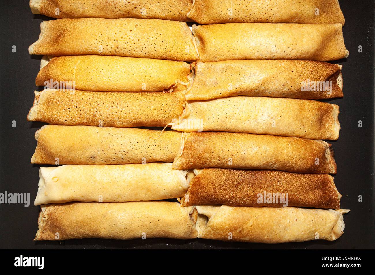 Pancake fritti e arrotolati impilati fianco a fianco su una superficie scura, vista dall'alto, primo piano Foto Stock