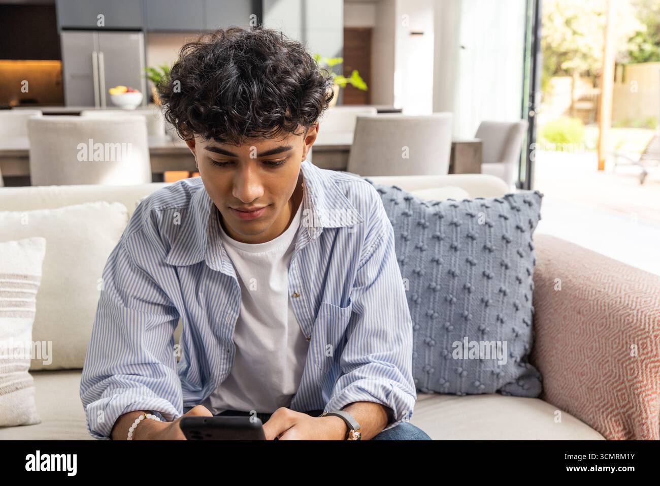 Uomo seduto sul divano che tiene lo smartphone accanto all'isola della cucina nel soggiorno Foto Stock