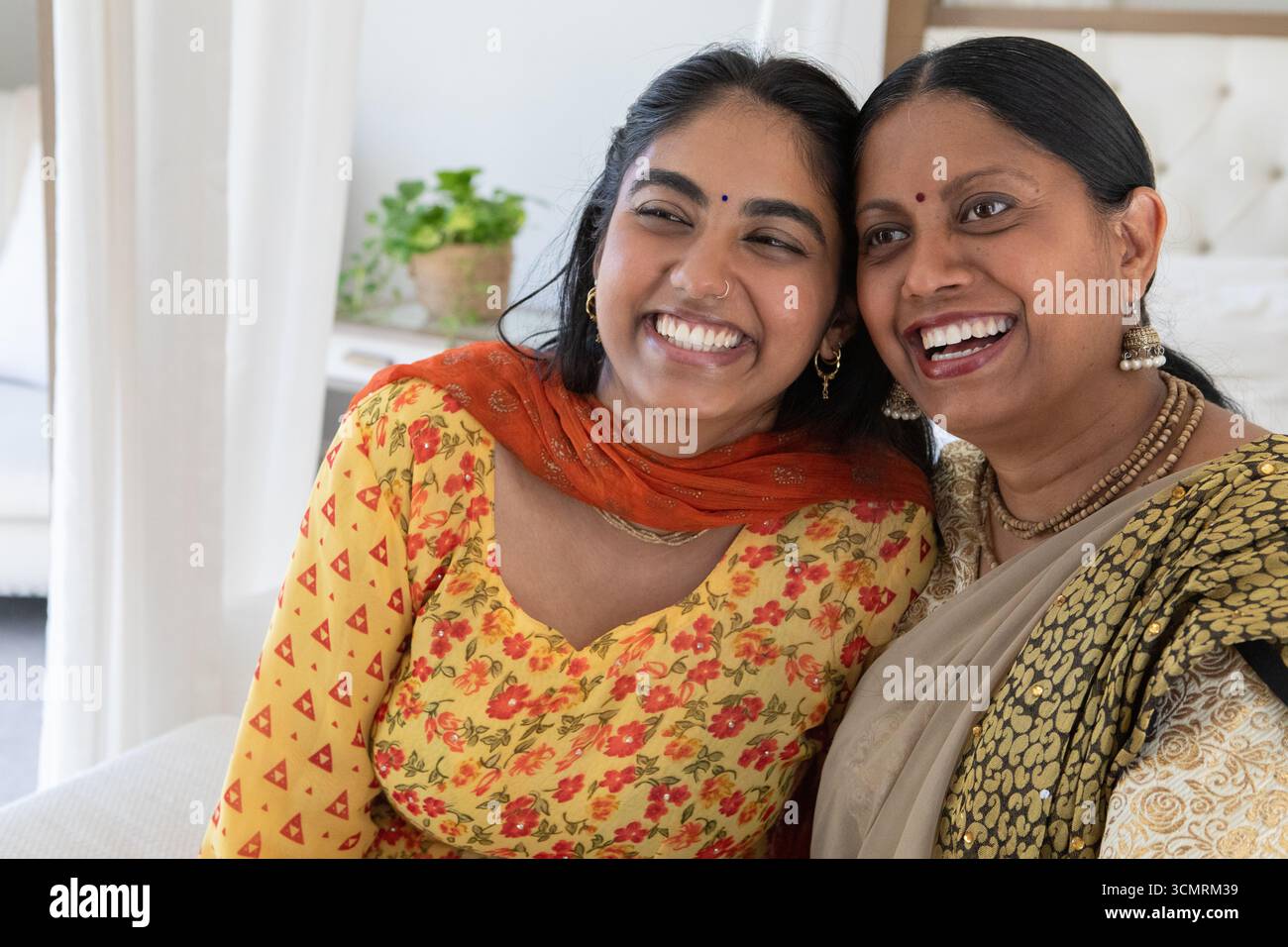 Madre indiana che indossa il sari e figlia nel salwar kameez sorride mentre siede sul divano a casa Foto Stock