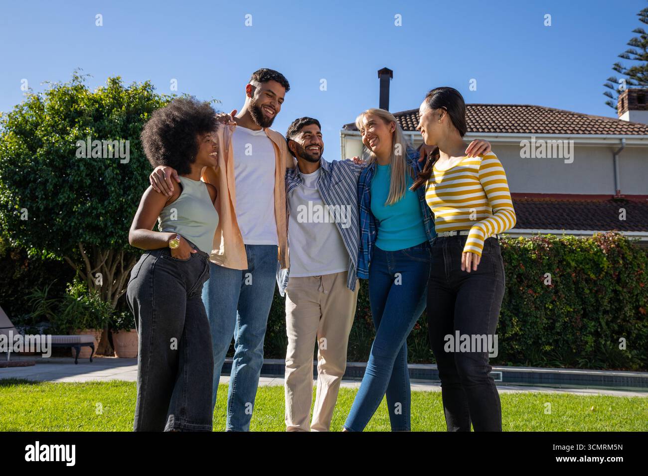 Amici diversi in piedi braccio su prato verde vicino alla piscina accanto alla siepe vicino alla casa Foto Stock