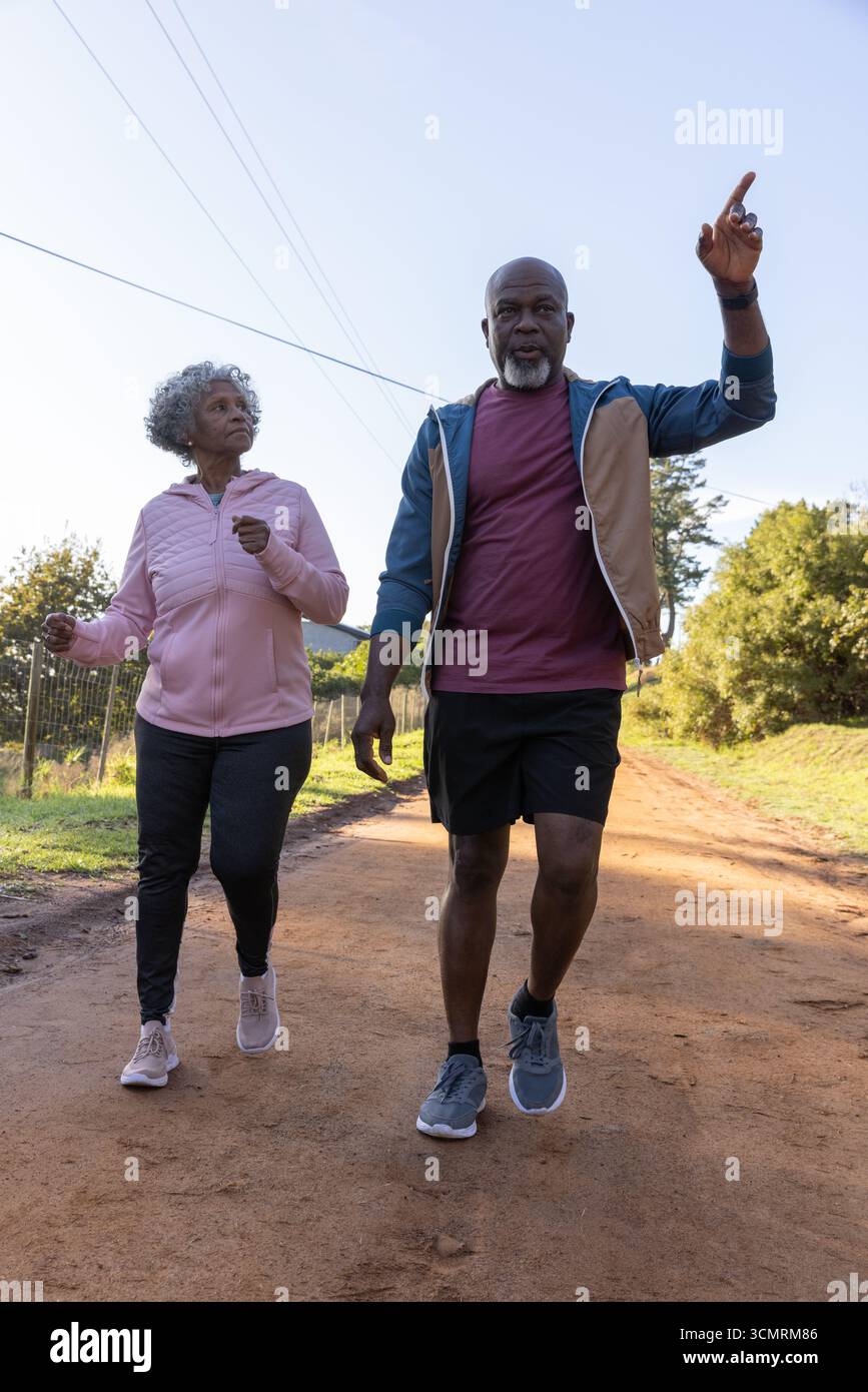 Coppia senior afroamericana che cammina su una strada sterrata che punta verso l'alto in felpa con cappuccio sotto le linee elettriche Foto Stock