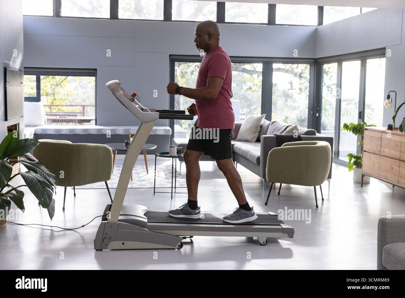 Senior afroamericano che cammina sul tapis roulant in camera e segue i gradini del fitness tracker Foto Stock Senior afroamericano che cammina sul tapis roulant in camera e segue i gradini del fitness tracker Foto Stock