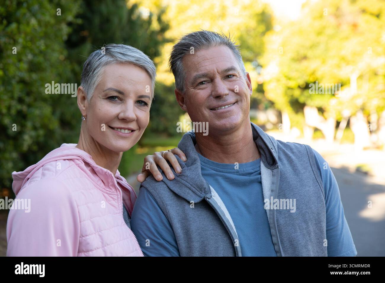 Coppia senior in piedi su un sentiero residenziale lastricato in giacche trapuntate e con zip sotto la luce del sole Foto Stock