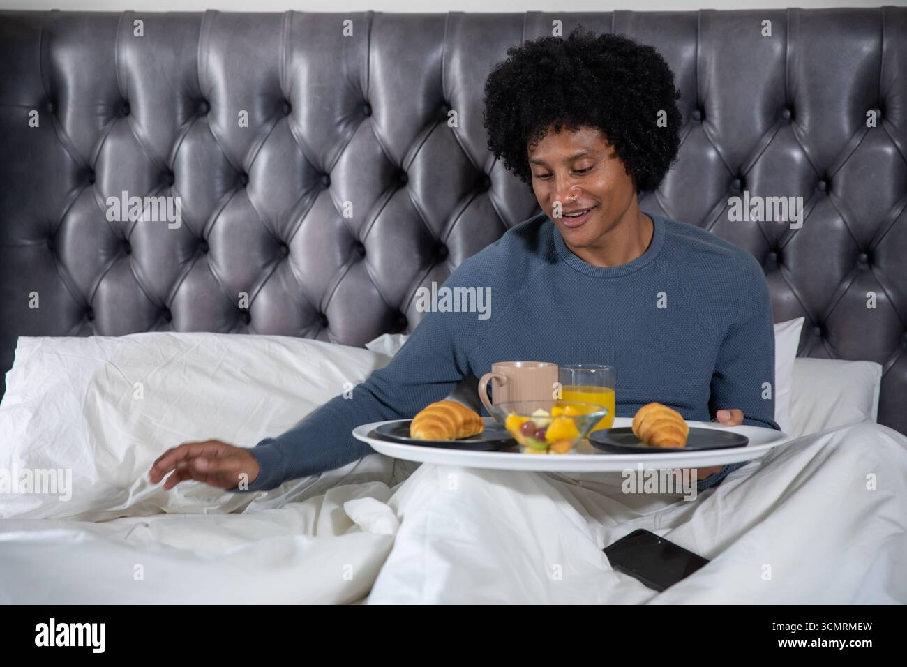 Uomo afroamericano medio adulto seduto sul letto con lenzuola adattate, vassoio per la colazione in camera da letto Foto Stock