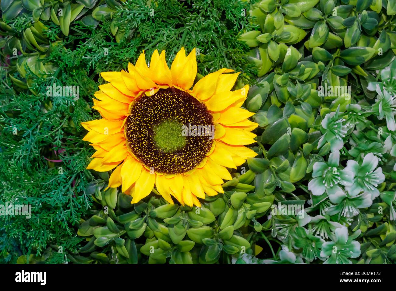 Un vibrante girasole artificiale con petali gialli si distingue per il verde decorativo in plastica, progettato come una realistica disposizione floreale per la casa Foto Stock