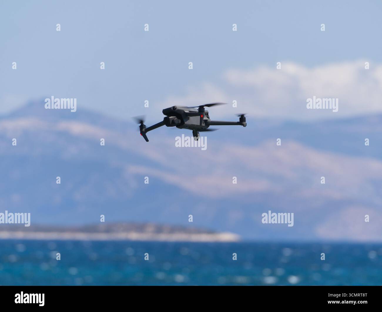 Drone in quadricottero nero che si libra nel cielo sopra l'acqua blu dell'oceano con montagne sullo sfondo Foto Stock