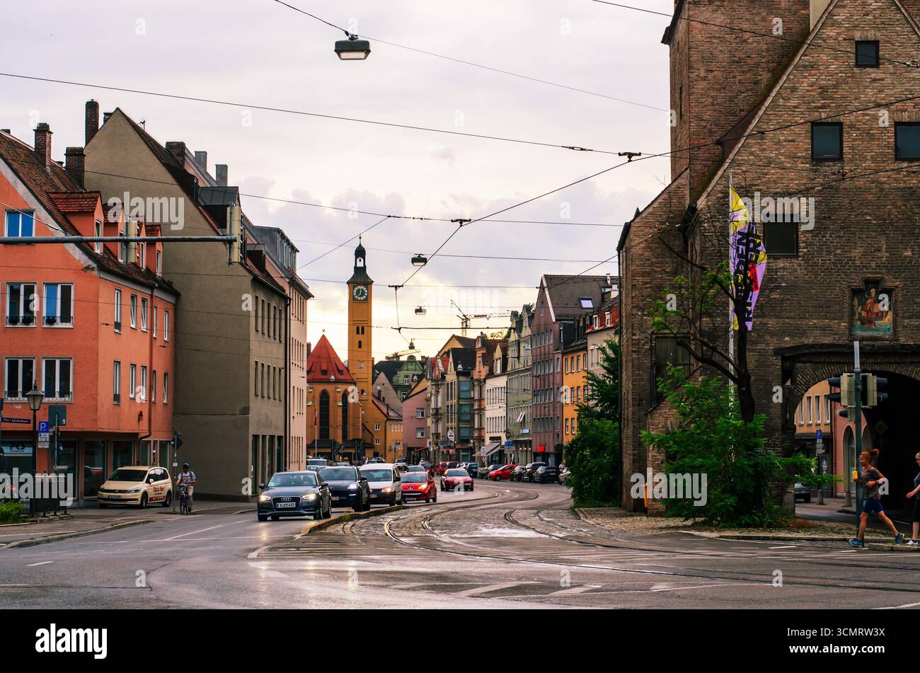AUGUSTA, GERMANIA - GIUGNO 27 2025: Strada della città con la storica torre dell'orologio sullo sfondo e persone che corrono lungo i marciapiedi durante la serata dorata Foto Stock