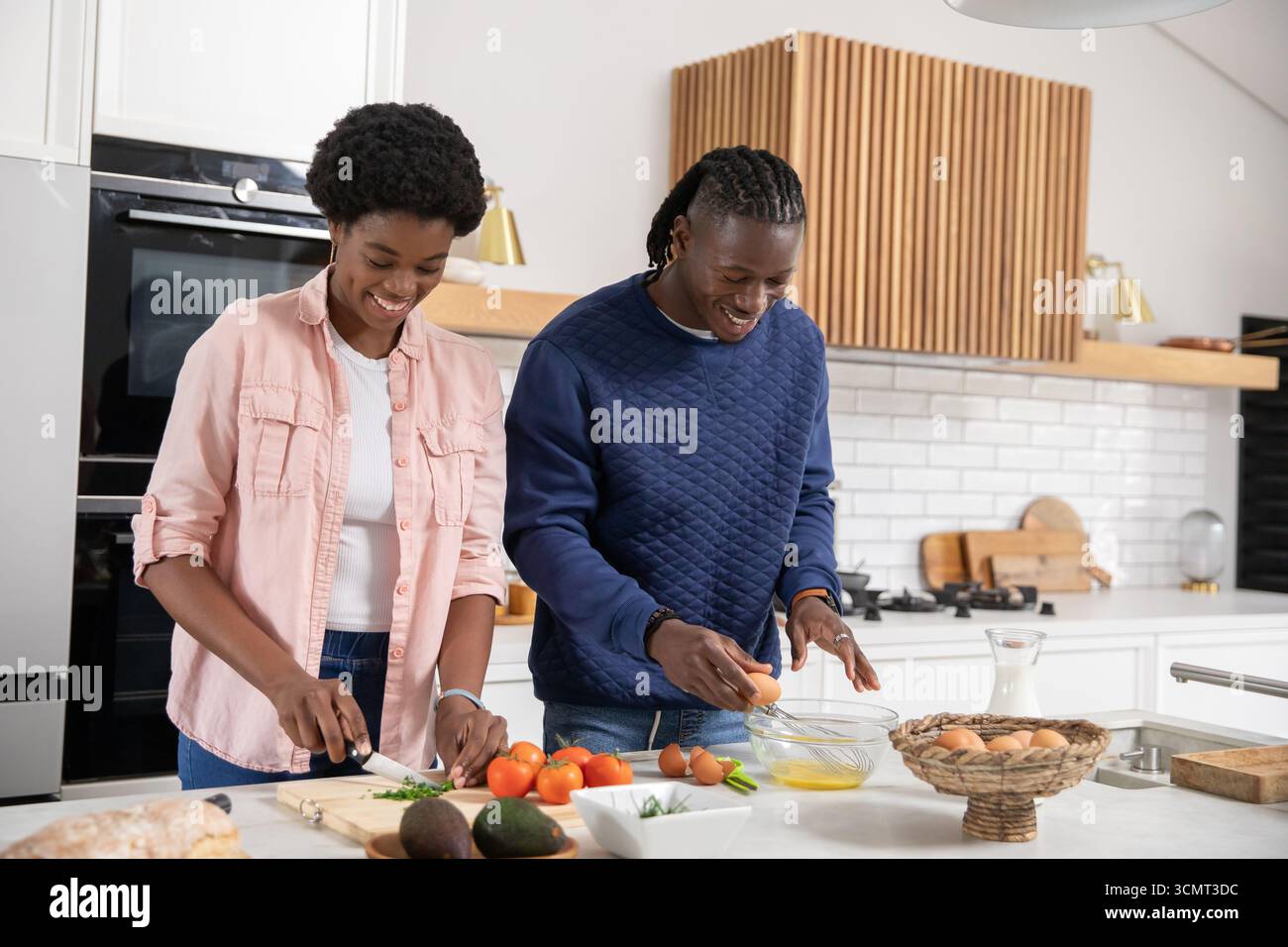 Coppia afroamericana che trita le erbe su una tavola di legno e che sbatte le uova nella ciotola in cucina Foto Stock
