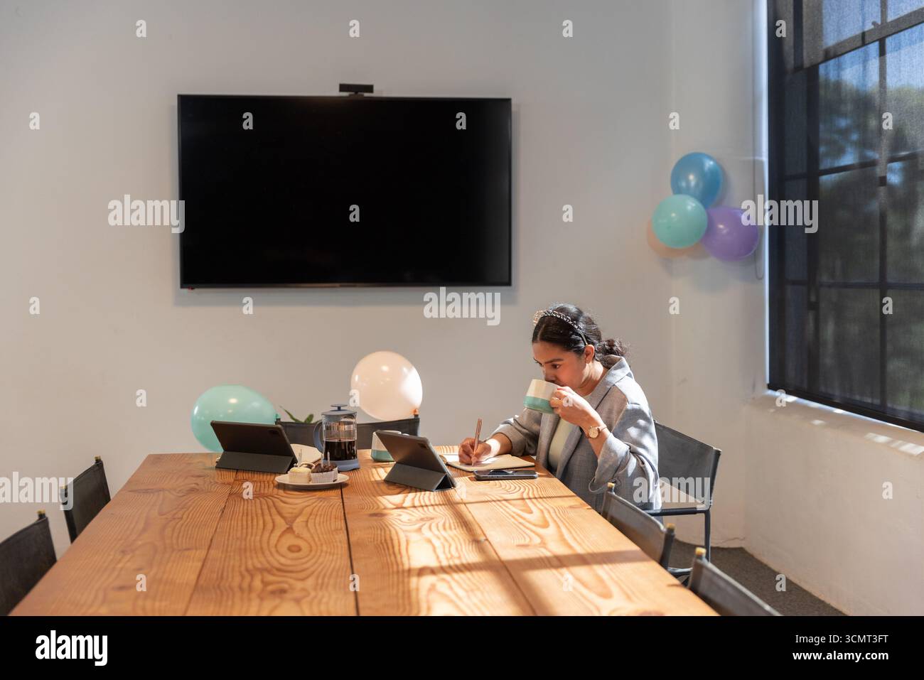 Donna asiatica che sorseggia caffè e prende appunti al tavolo delle conferenze, tablet, palloncini, spazio per copiare Foto Stock